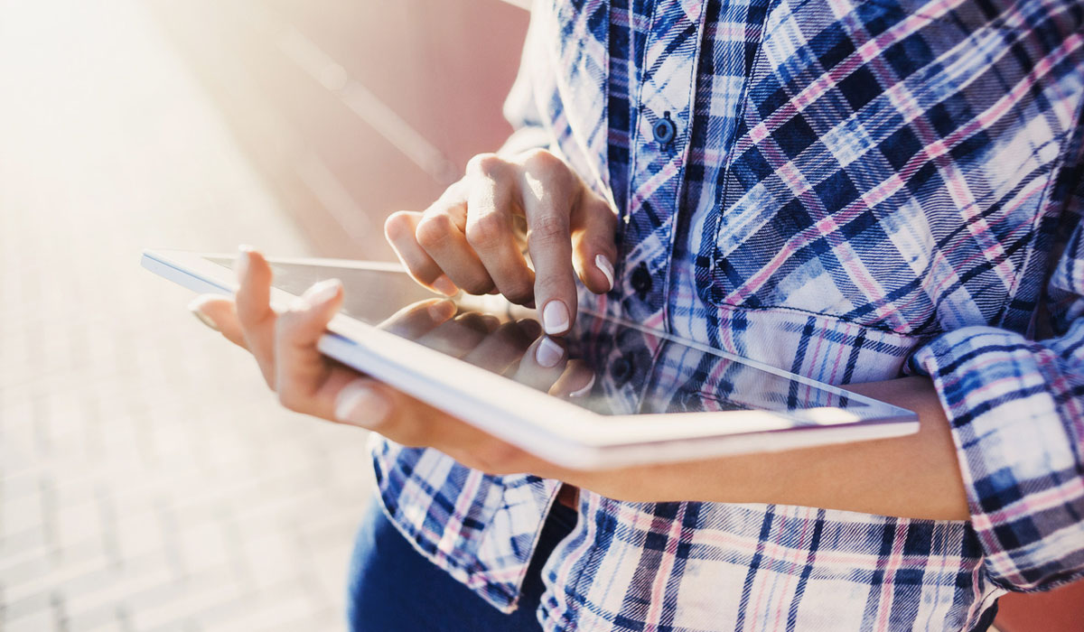 A person using a tablet outdoors, focusing on the screen with one hand.