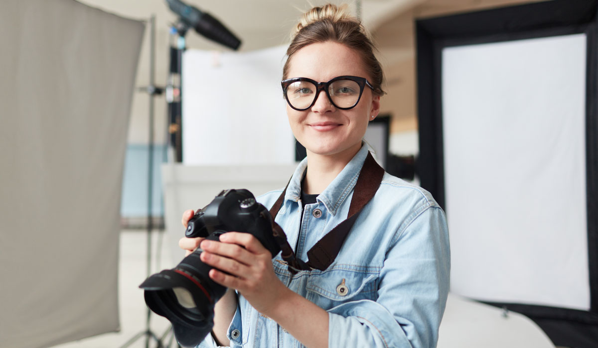 A smiling woman wearing glasses holds a camera in a photography studio setting.