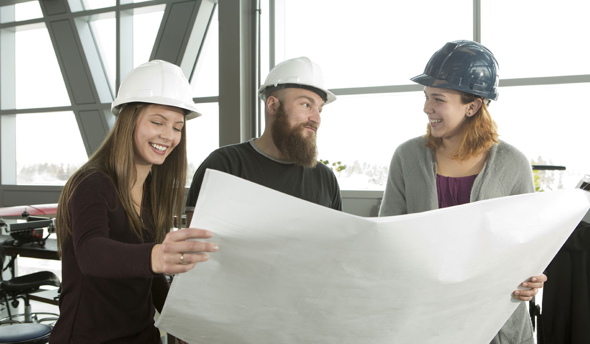 Three construction professionals reviewing blueprints together, wearing hard hats and smiling in a modern workspace.