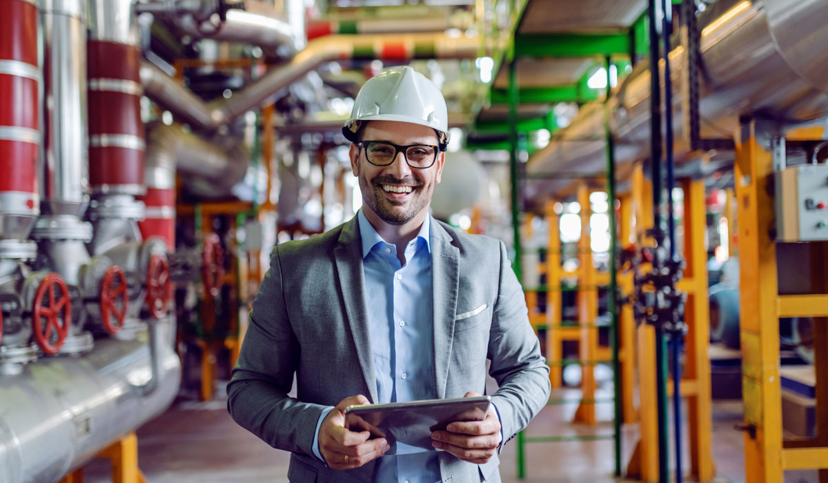 A professional in a hard hat and suit smiling while holding a tablet in an industrial setting.