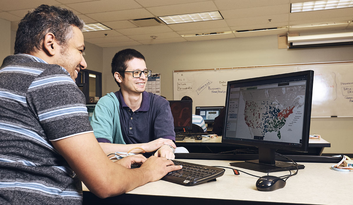 Two men collaborate at a computer, analyzing a colorful map of the United States displayed on the screen in an indoor setting.