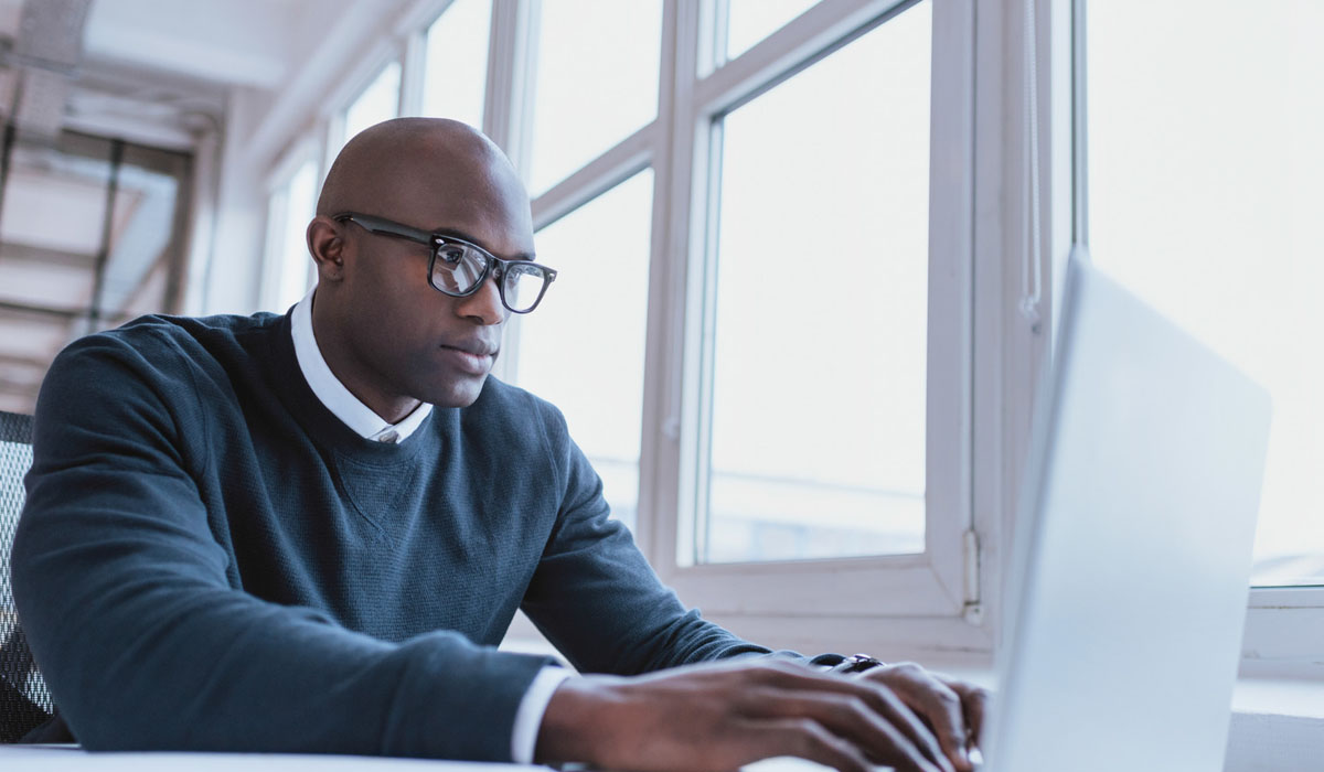A focused man wearing glasses works intently on a laptop by a large window.