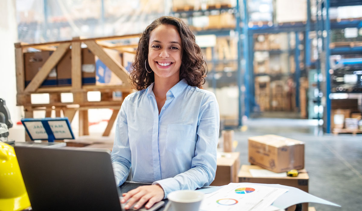 A woman smiling while working on a laptop in a warehouse setting, with charts and boxes in the background.