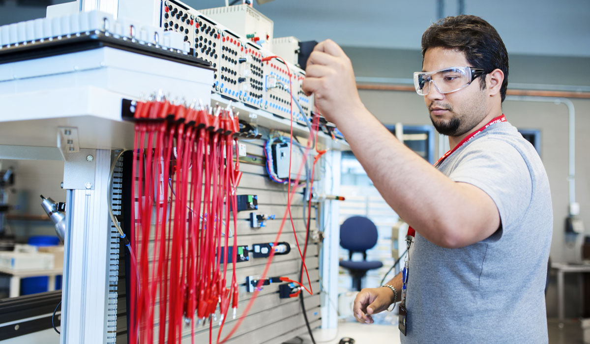 A student wearing safety goggles is working on an electrical circuit setup in a lab environment.