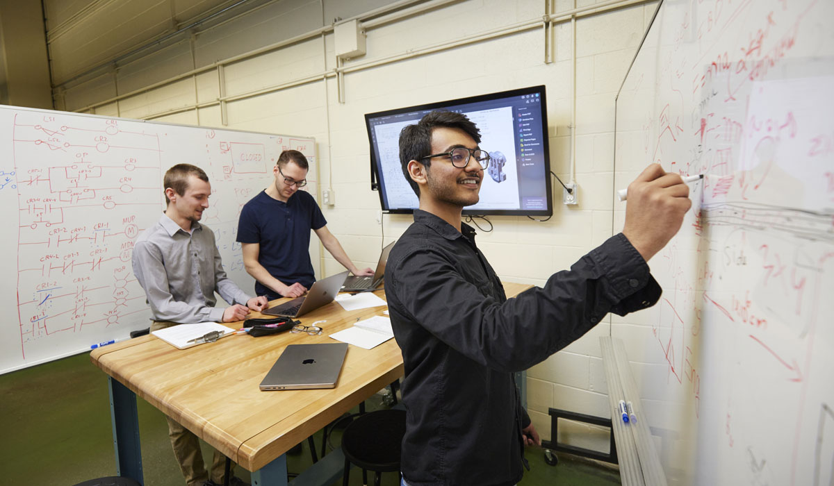 Three students collaborate on a project in a modern workspace, with one writing on a whiteboard while the others work on laptops.