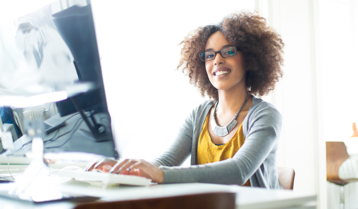 A smiling woman with curly hair working at a desktop computer, wearing glasses and a stylish necklace.