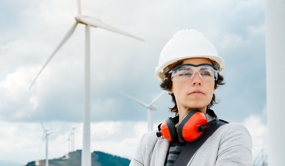 A woman wearing a hard hat and protective gear stands confidently in front of wind turbines.