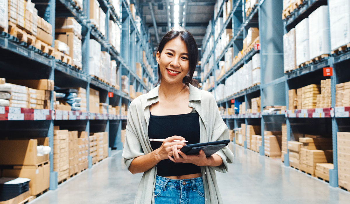 A smiling woman holding a tablet stands in a well-organized warehouse aisle.