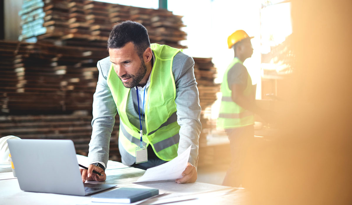 A man in a safety vest working on a laptop in a construction environment, while another worker in the background wears a hard hat.