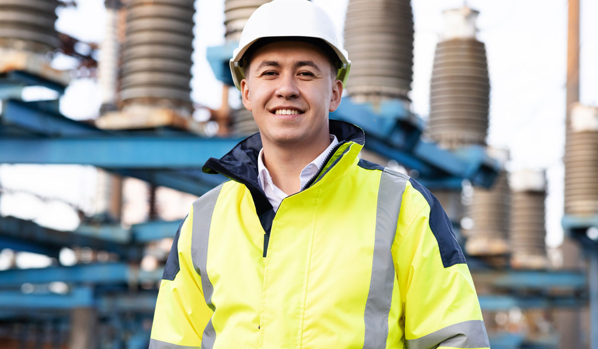 A smiling man wearing a high-visibility jacket and hard hat, standing in front of industrial equipment.