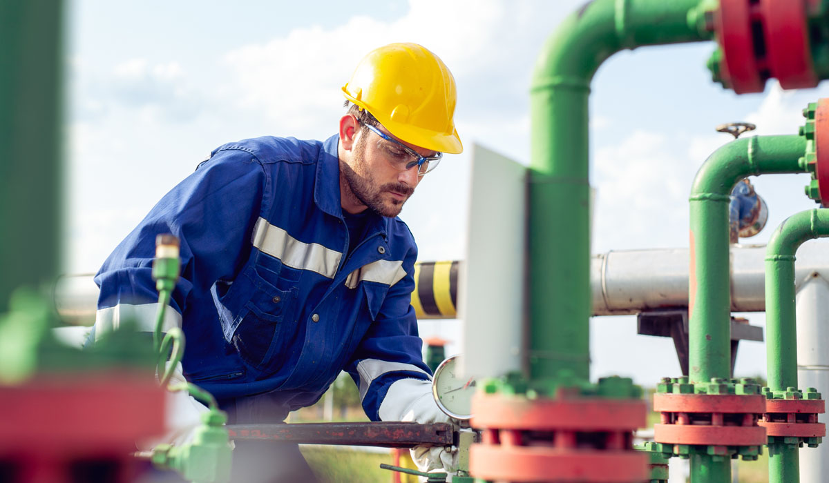 A worker in a yellow hard hat and safety glasses is inspecting machinery with valves and pipes in an industrial setting.