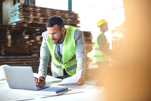 a man leaning over a desk reaching toward a computer, wearing a high visbility vest on a construction site.