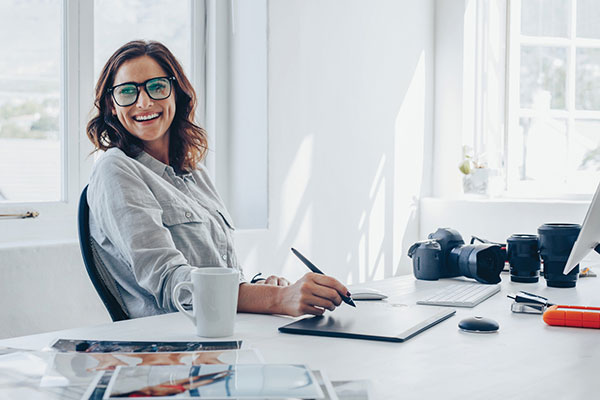 A woman works on a tablet with her stylus. Professional camera equipment is on the desk beside her.