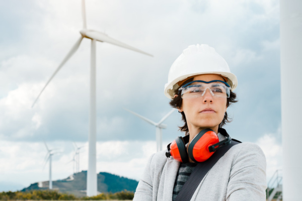 a woman with a serious expressions staring into the distance, wearing construction equipment near a wind turbine.