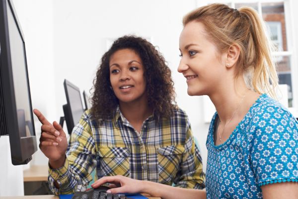 Two smiling ladies stand side by side, looking at a computer screen. One is instructing the other.