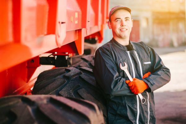 A service technician holds a wrench and leans against a tractor wheel.