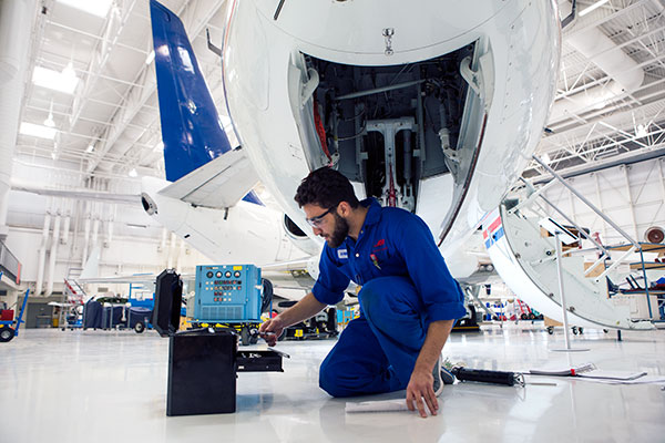 A male student kneels beside a tool box. He's wearing safety glasses and coveralls. An airplane engine is open behind him. 