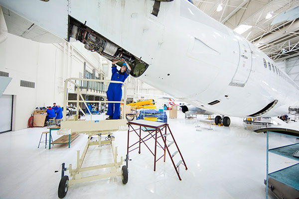 A woman wearing safety glasses and coveralls works on the engine under an airplane.