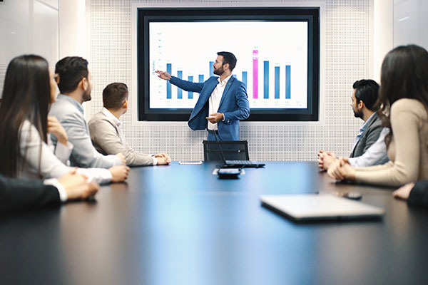 A manager stands at the front of a board room, explaining data seen on the smart board. His team sits around a table listening.