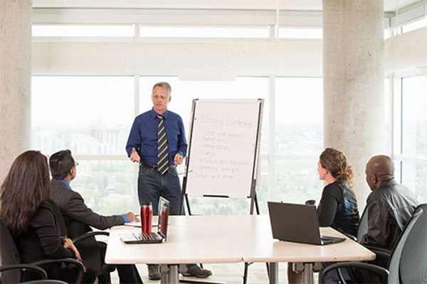 A man wearing a blue button-down shirt and tie, presents to a group of four professional seated around a table. 