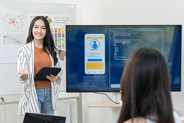 A woman holding an ipad with wireframes displayed on a monitor beside her