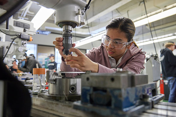 A young woman wearing safety glasses secures a drillbit as she prepares to use a drill on metalwork.