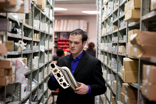 An apprentice stands between shelves in a warehouse looking at a part.