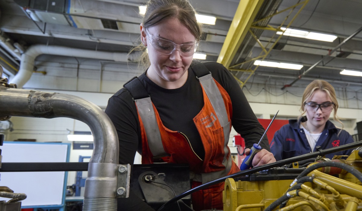 “Two people wearing safety glasses work on a large piece of machinery in a workshop; one is using a screwdriver while the other observes, surrounded by industrial equipment and tools.
