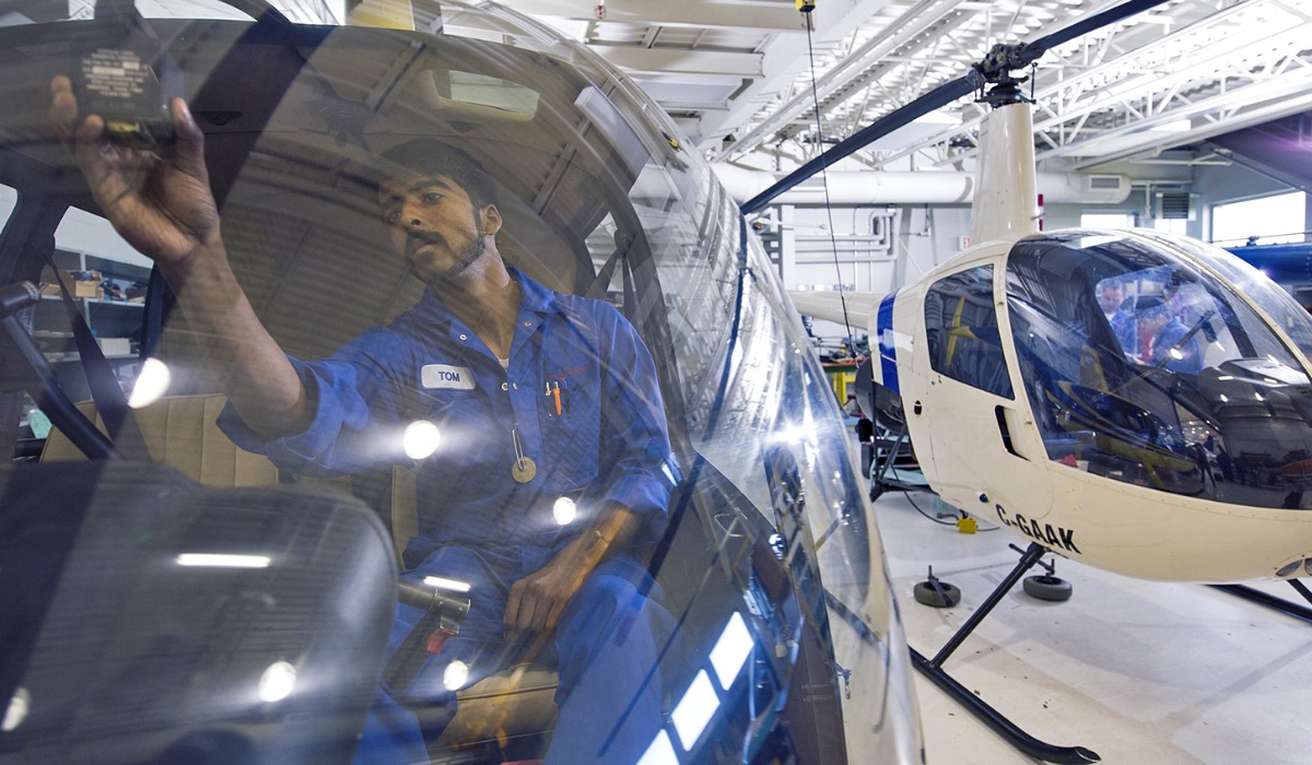 A technician performing maintenance on a helicopter, with another helicopter visible in the background.