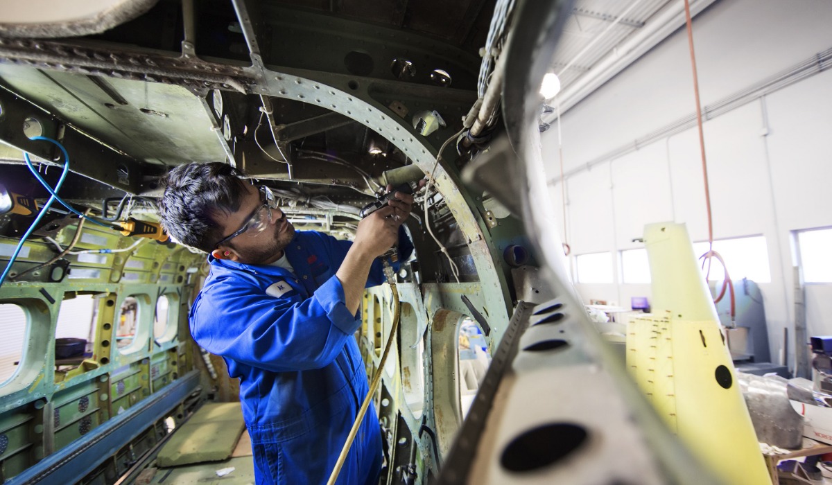 A technician in a blue jumpsuit works on the interior structure of an aircraft, focusing on wiring and maintenance tasks.