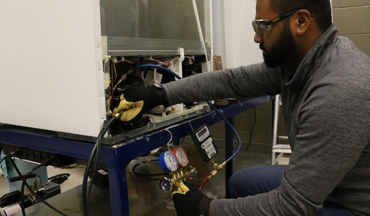 A technician wearing safety glasses and gloves uses pressure gauges and tools to service the internal components of an appliance on a workbench.