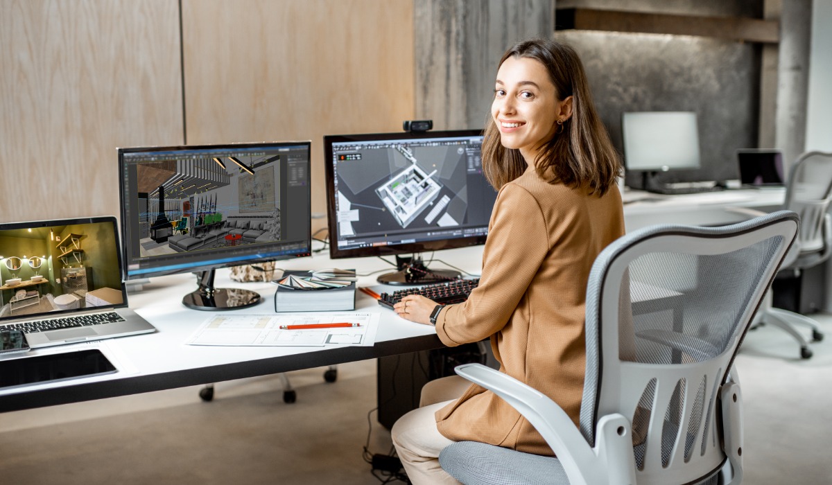 A smiling woman sits at a modern workspace with dual computer monitors displaying architectural designs.