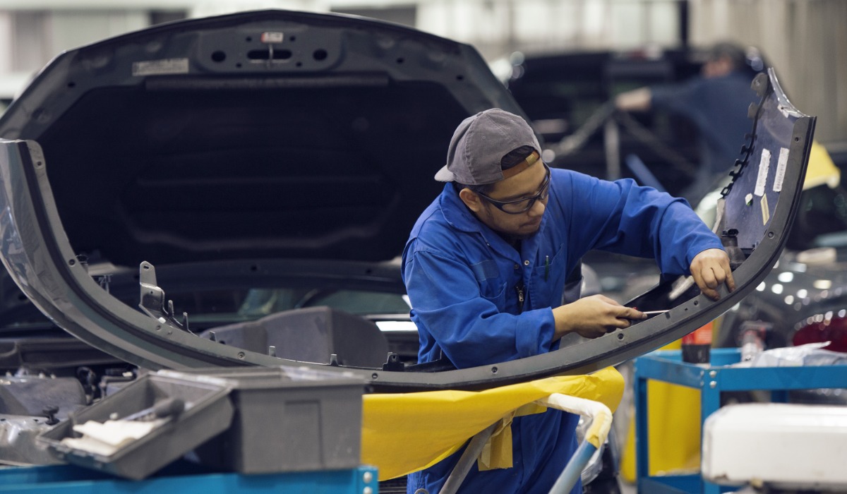 A student wearing safety glasses, a blue coverall, and a backward cap repairs a car bumper in an auto body shop, surrounded by tools and equipment.