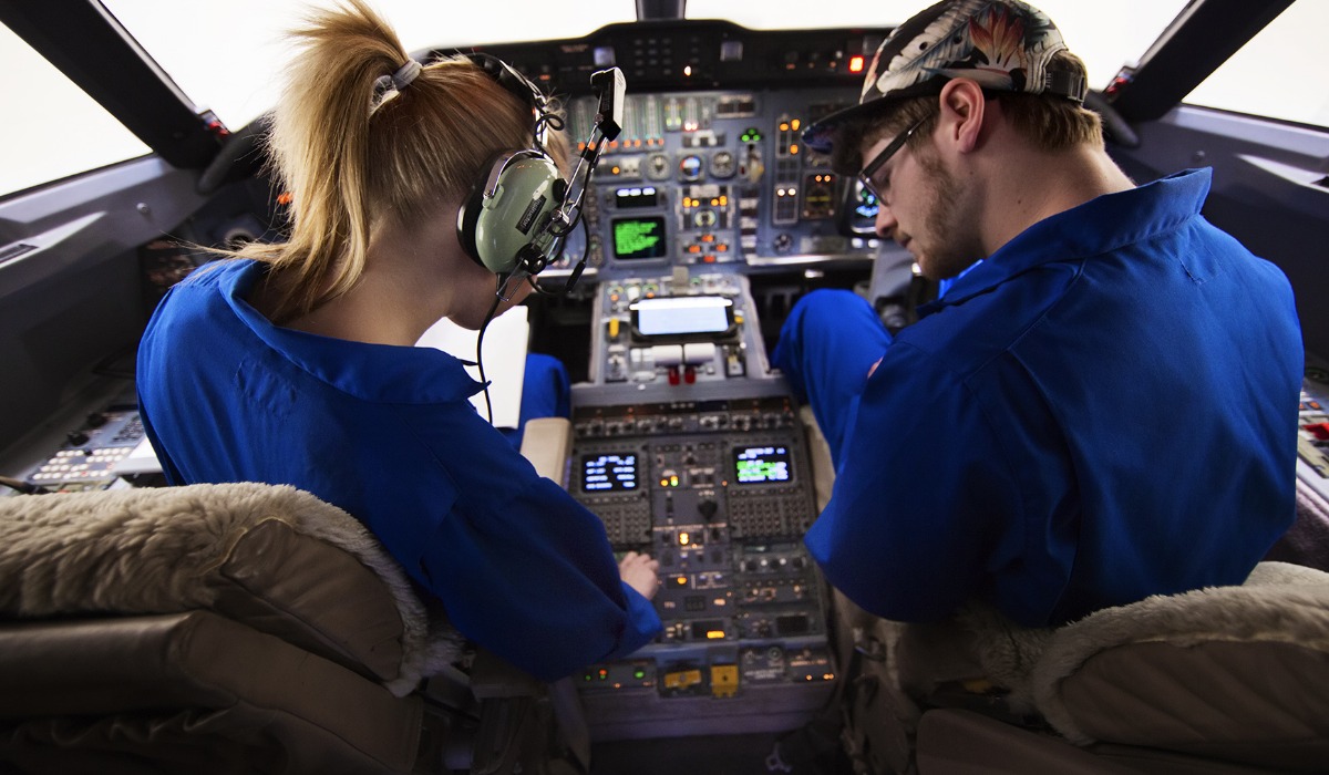 Two students in blue uniforms are seated in a cockpit, focused on the control panel and instruments.