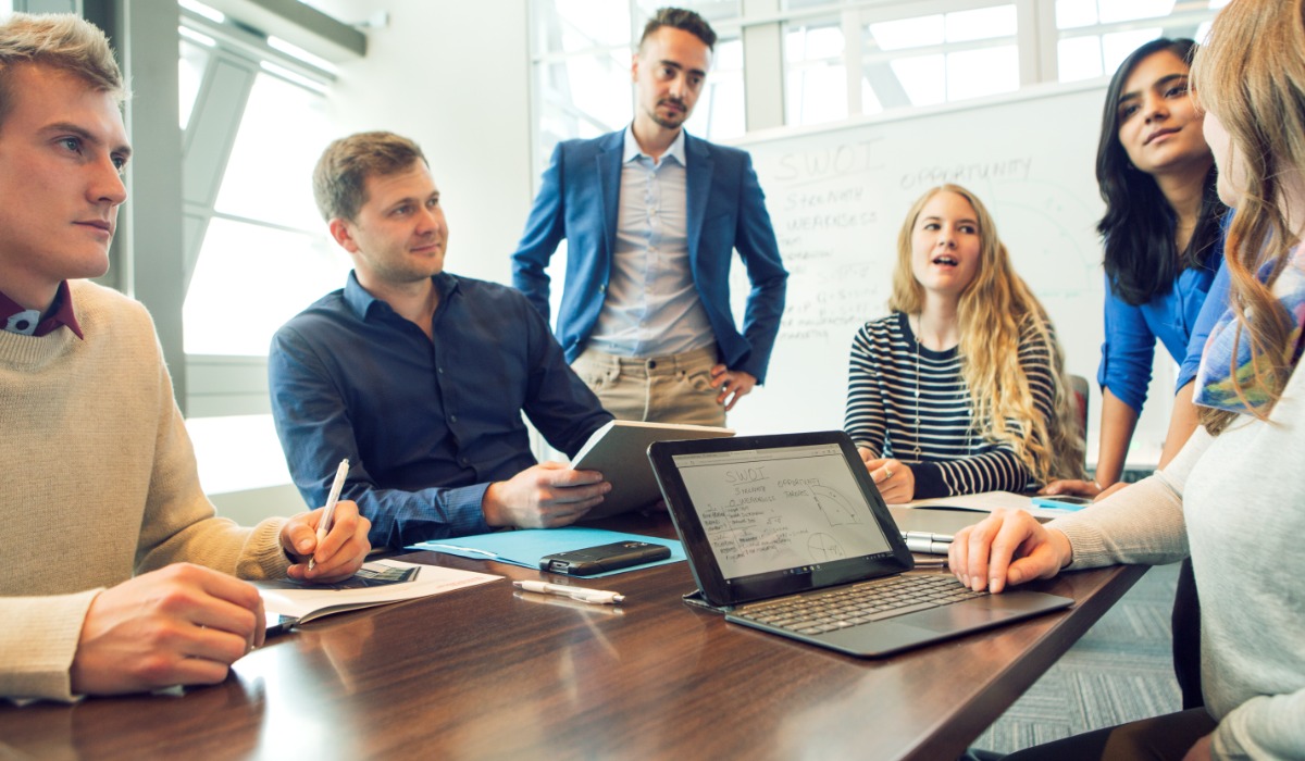 A diverse group of professionals engaged in a collaborative discussion around a conference table, working on a project with laptops and notebooks.