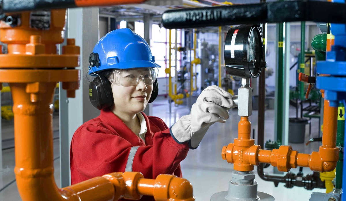 A technician wearing a blue helmet and gloves is working on industrial piping in a facility, adjusting equipment with precision.