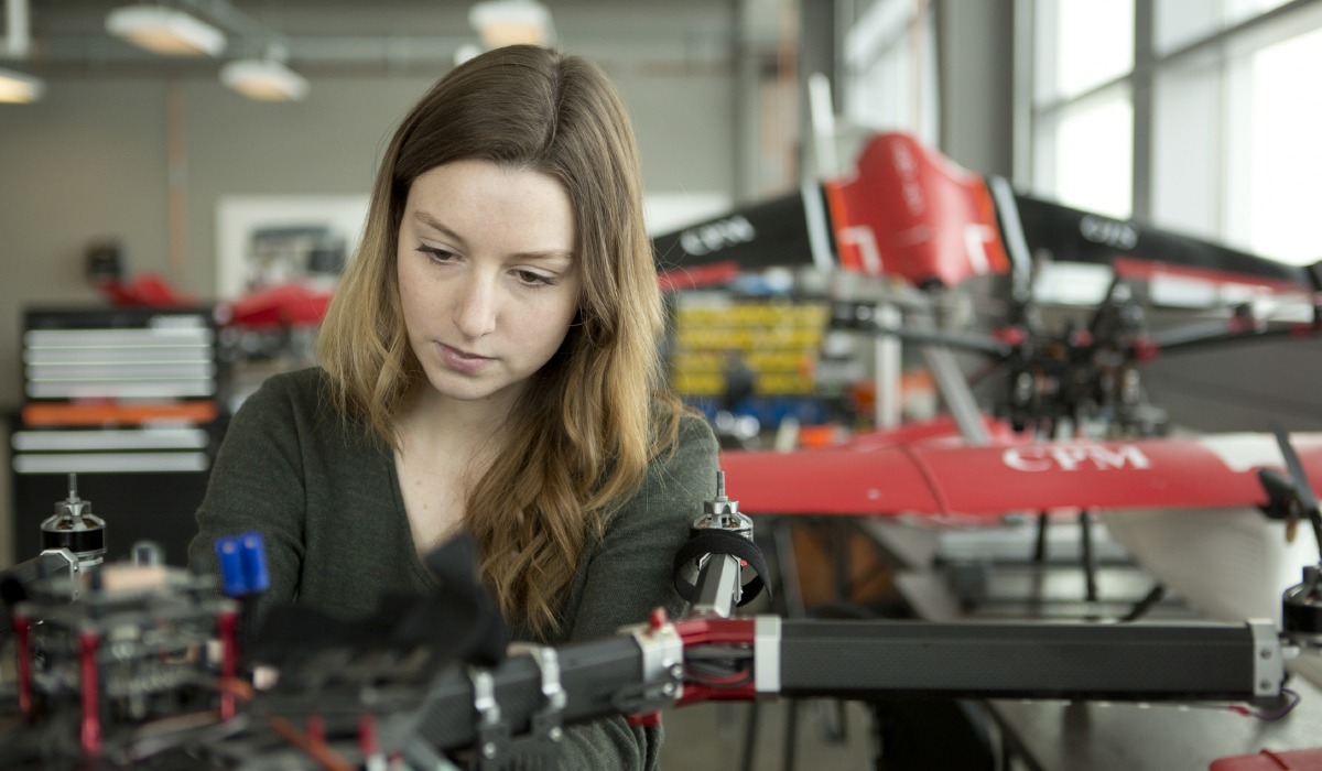 A woman intently working on a drone in a well-equipped workshop, with another drone visible in the background.