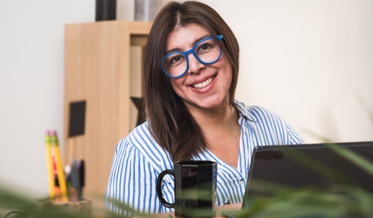 A smiling woman wearing blue glasses sits at her desk with a laptop, a black mug, and colorful pens in the background.