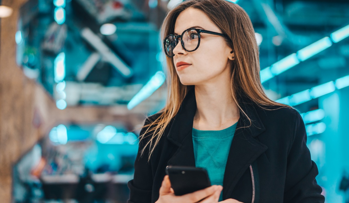 A young woman with glasses holding a smartphone, looking thoughtfully in an urban environment with neon lighting.
