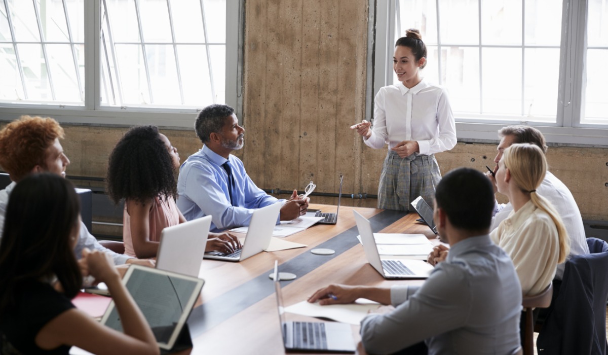 A professional woman giving a presentation to a diverse group of colleagues seated around a conference table with laptops.
