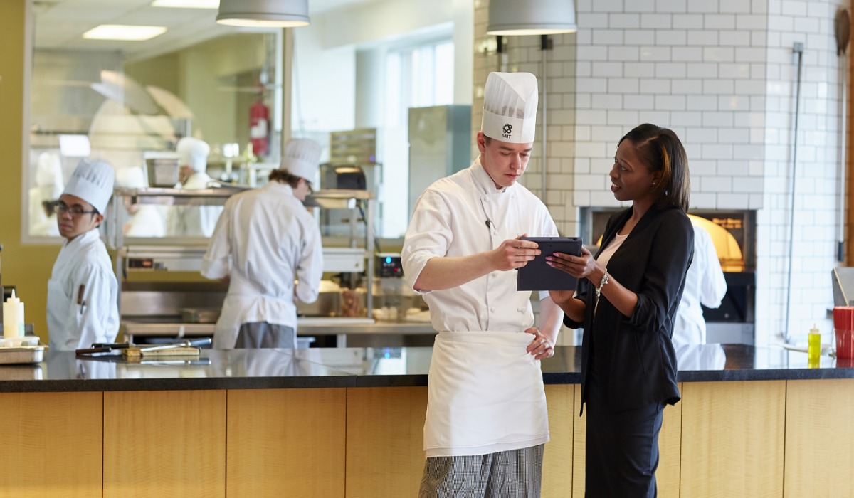A chef and a businesswoman discuss on a tablet in a modern kitchen, with other chefs working in the background.
