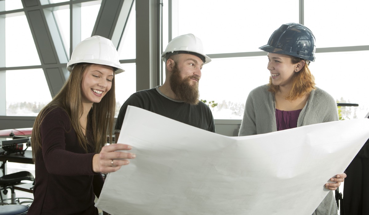 Three construction professionals, two wearing white hard hats and one wearing a blue hard hat, collaborate while reviewing a large blueprint inside a bright, modern workspace.