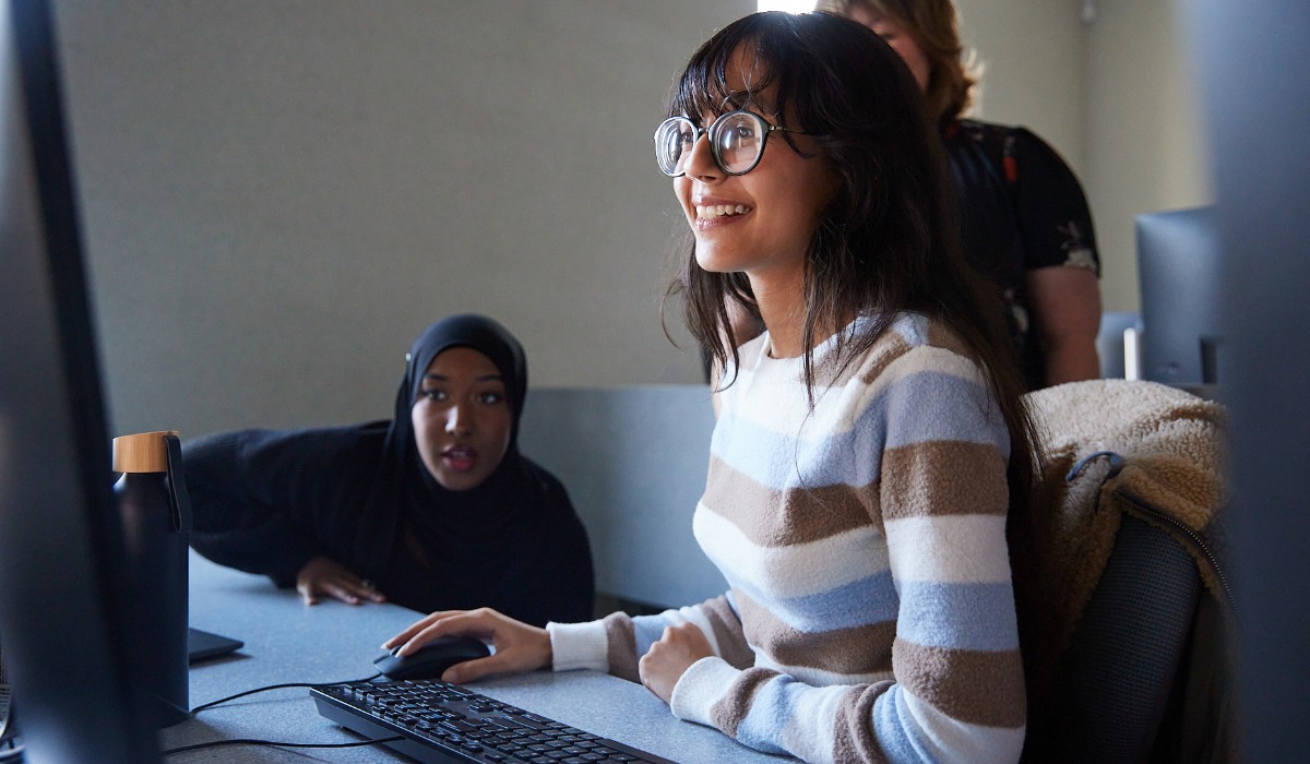 A young woman with glasses smiles while using a computer, with a surprised friend in a hijab watching her from behind.