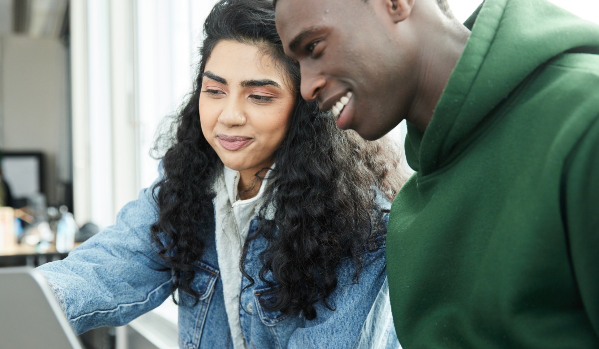Two young adults, a woman with curly hair and a man, are collaborating on a laptop while smiling at each other in a bright indoor setting.