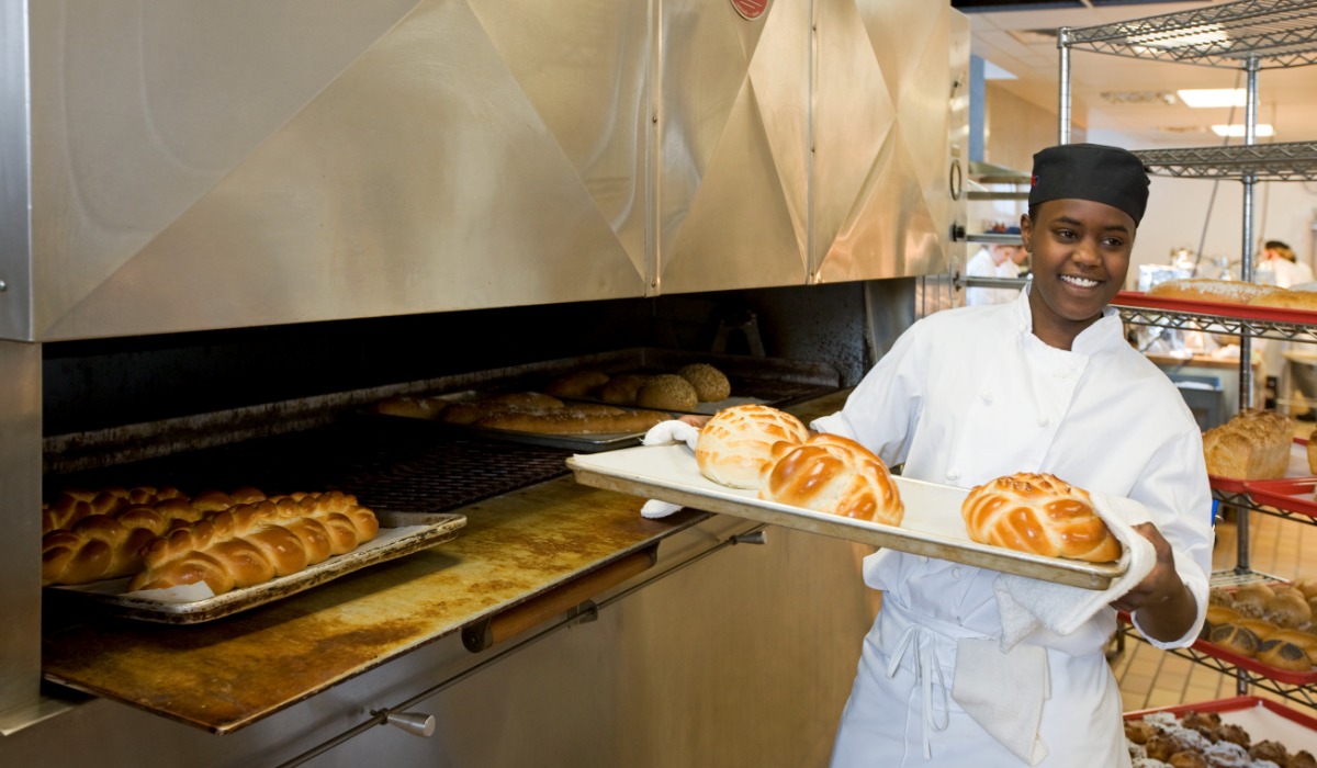 A baker proudly holds a tray of freshly baked bread rolls, with an oven in the background.