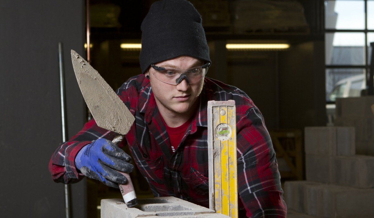 A bricklayer wearing safety glasses, gloves, and a beanie carefully applies mortar with a trowel while using a level to align concrete blocks.