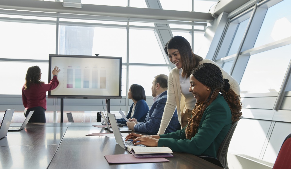 A business meeting in a modern office setting, featuring team members collaborating around a table while a presentation is displayed on a screen.