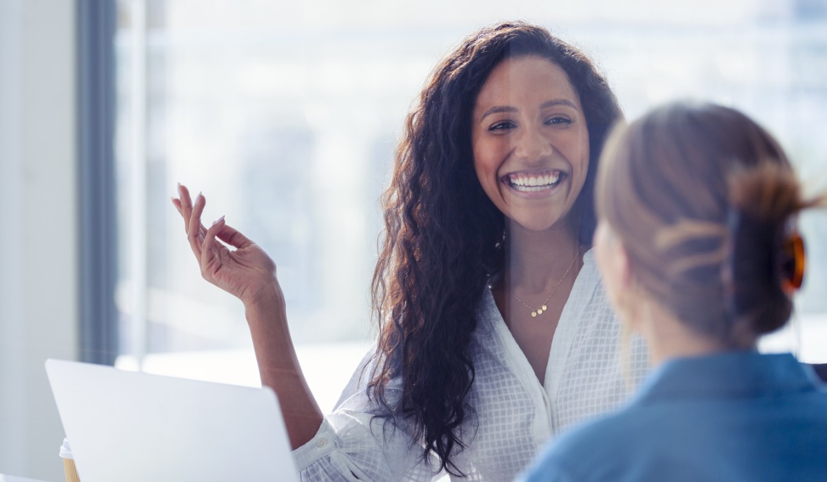 A woman with curly hair smiles while engaging in conversation with another person. The setting suggests a professional environment.