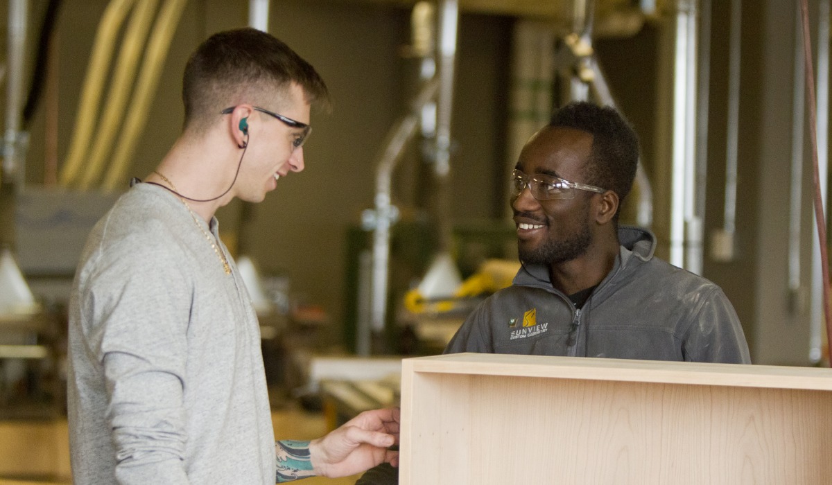 Two people wearing safety glasses smile and discuss a wooden cabinet project in a workshop with woodworking tools and equipment in the background.