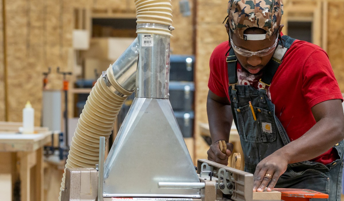 “A person wearing safety glasses, overalls, and a camouflage cap carefully operates a woodworking machine in a carpentry workshop.